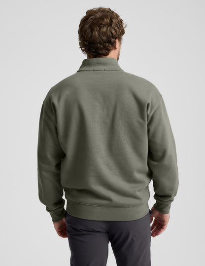 Man wearing a green quarter zip pullover and khaki pants with a plain background.