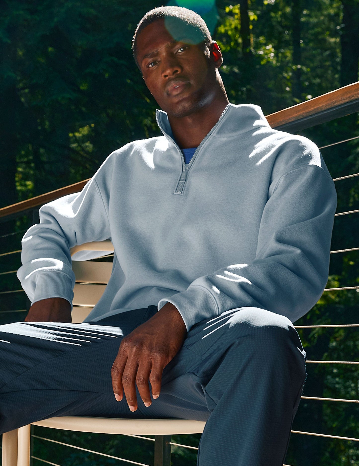 Man wearing a light blue pullover and dark pants sitting outdoors with greenery in the background.