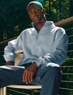 Man wearing a light blue pullover and dark pants sitting outdoors with greenery in the background.