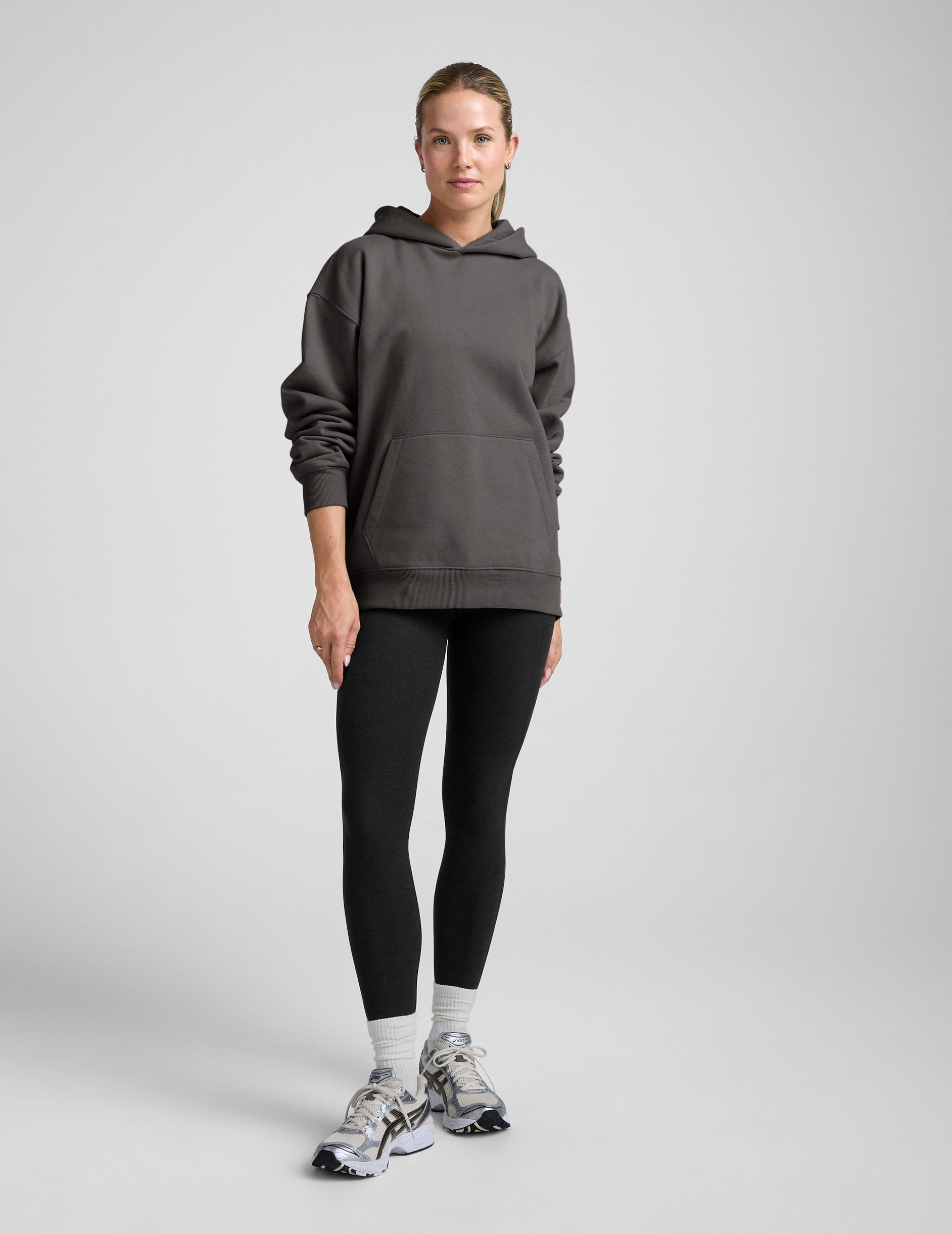 woman wearing a black hoodie with black leggings on a plain background.