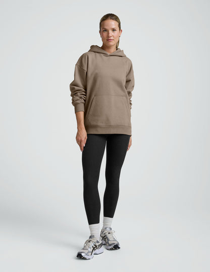 woman wearing a beige hoodie and black leggings on a plain background.
