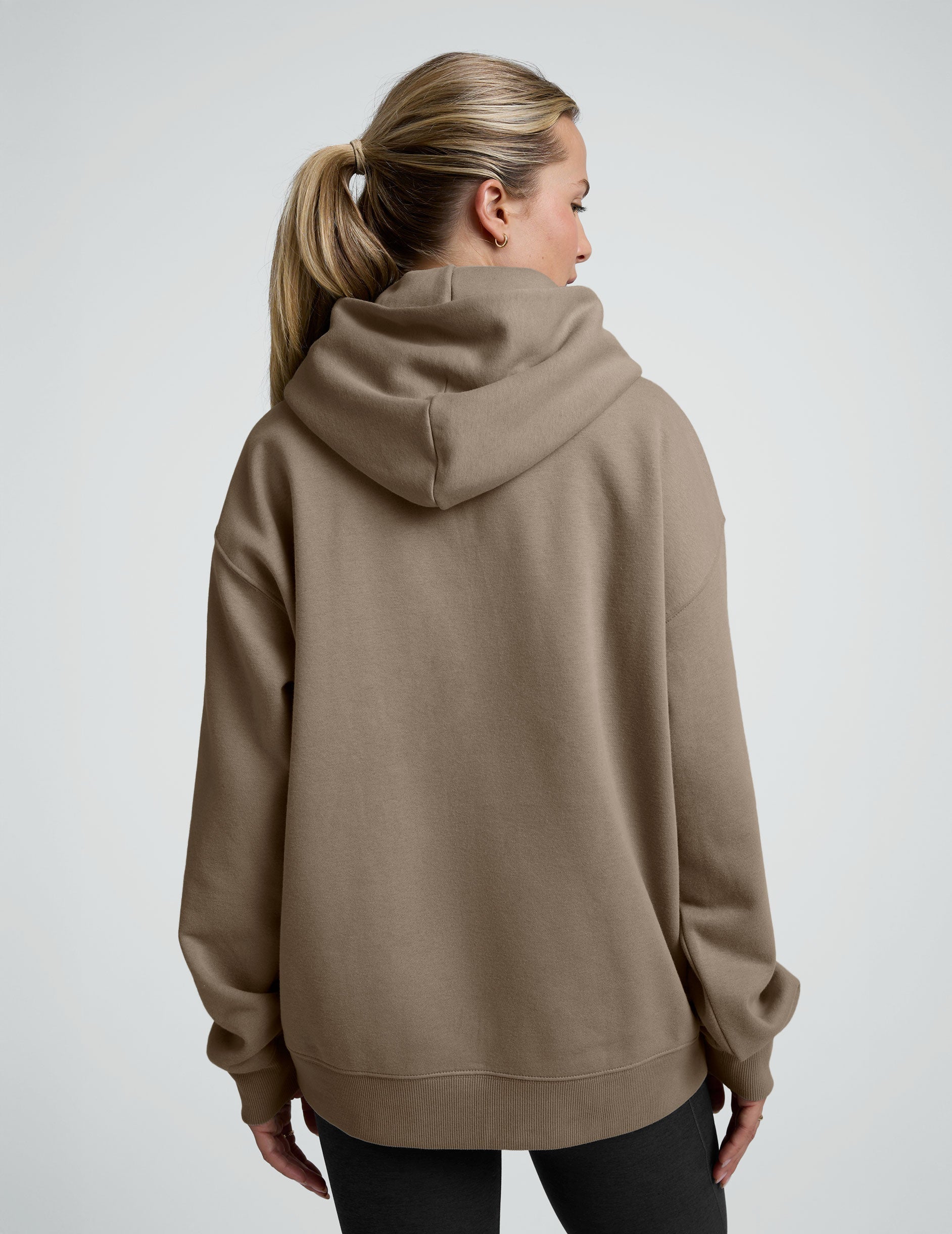 woman wearing a beige hoodie and black leggings on a plain background.