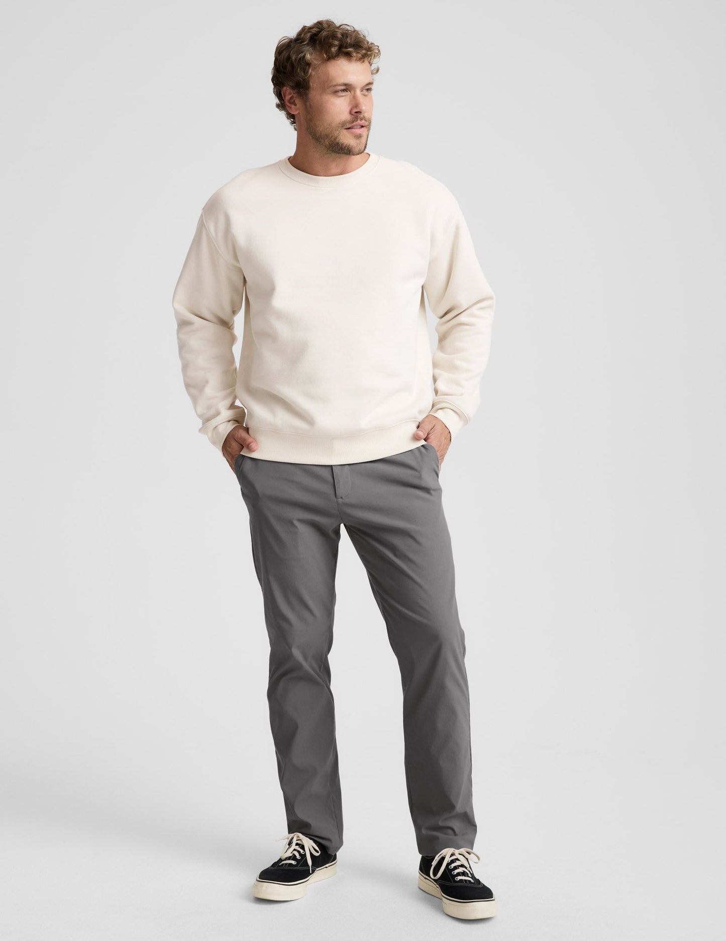 Man wearing a beige sweater and gray pants on a plain background.