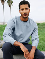 Man wearing a light blue sweatshirt sitting outdoors with palm trees in the background.