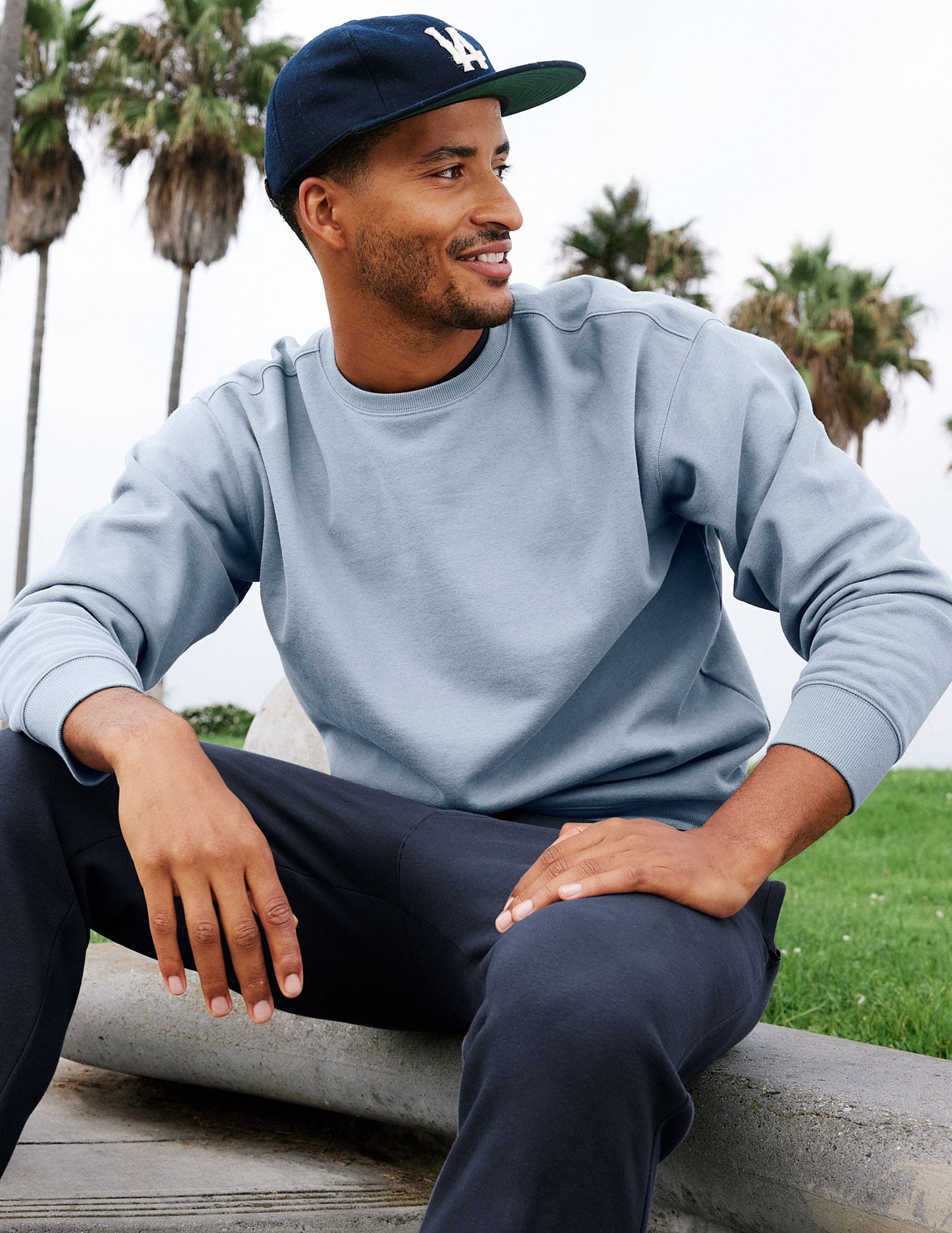 Man wearing a light blue sweatshirt and navy pants sitting outdoors with palm trees in the background.