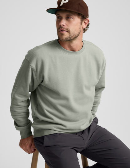Man wearing a green crewneck sweatshirt and dark pants sitting on a stool against a light gray background.