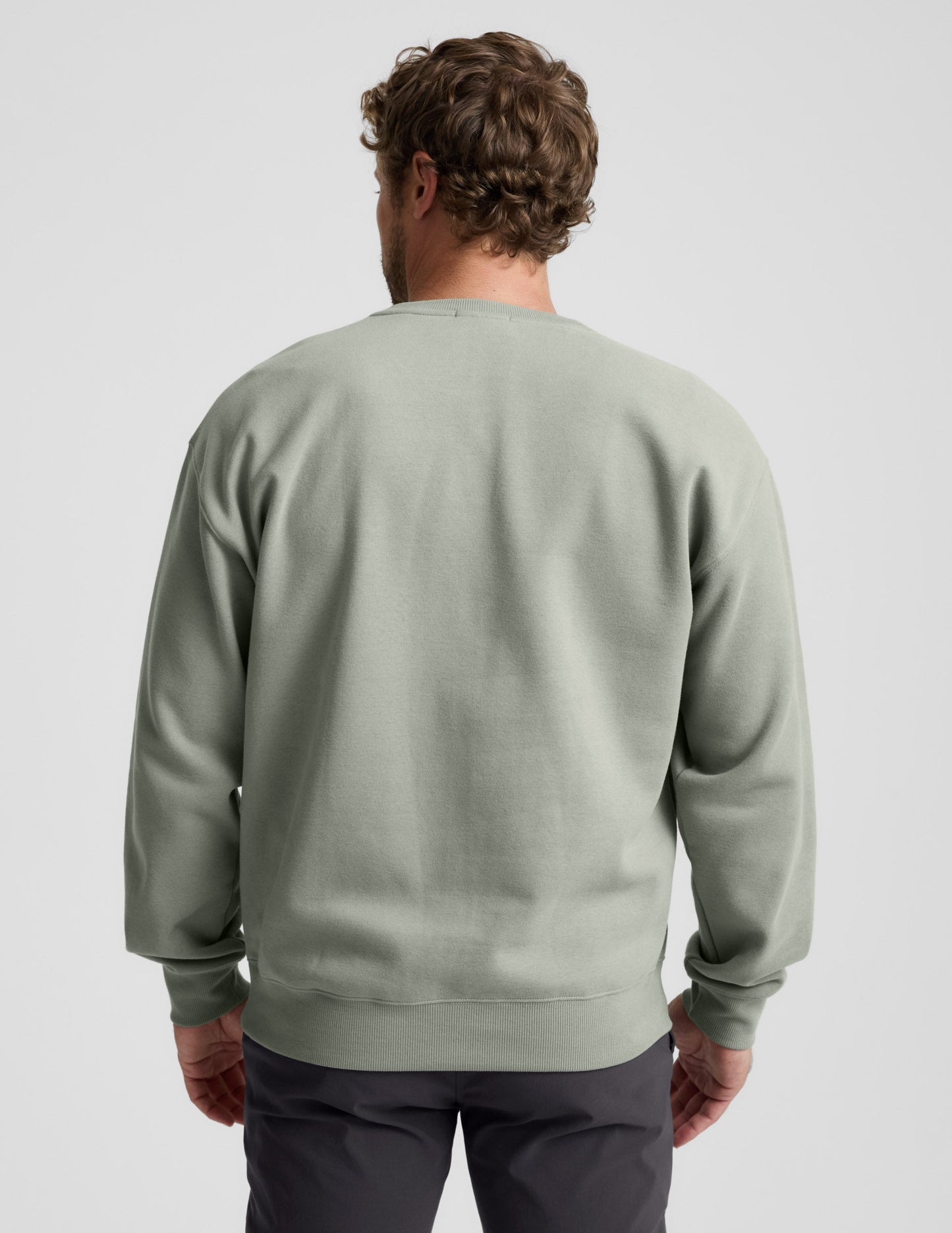 Man wearing a green crewneck sweatshirt and dark pants on a light gray background.