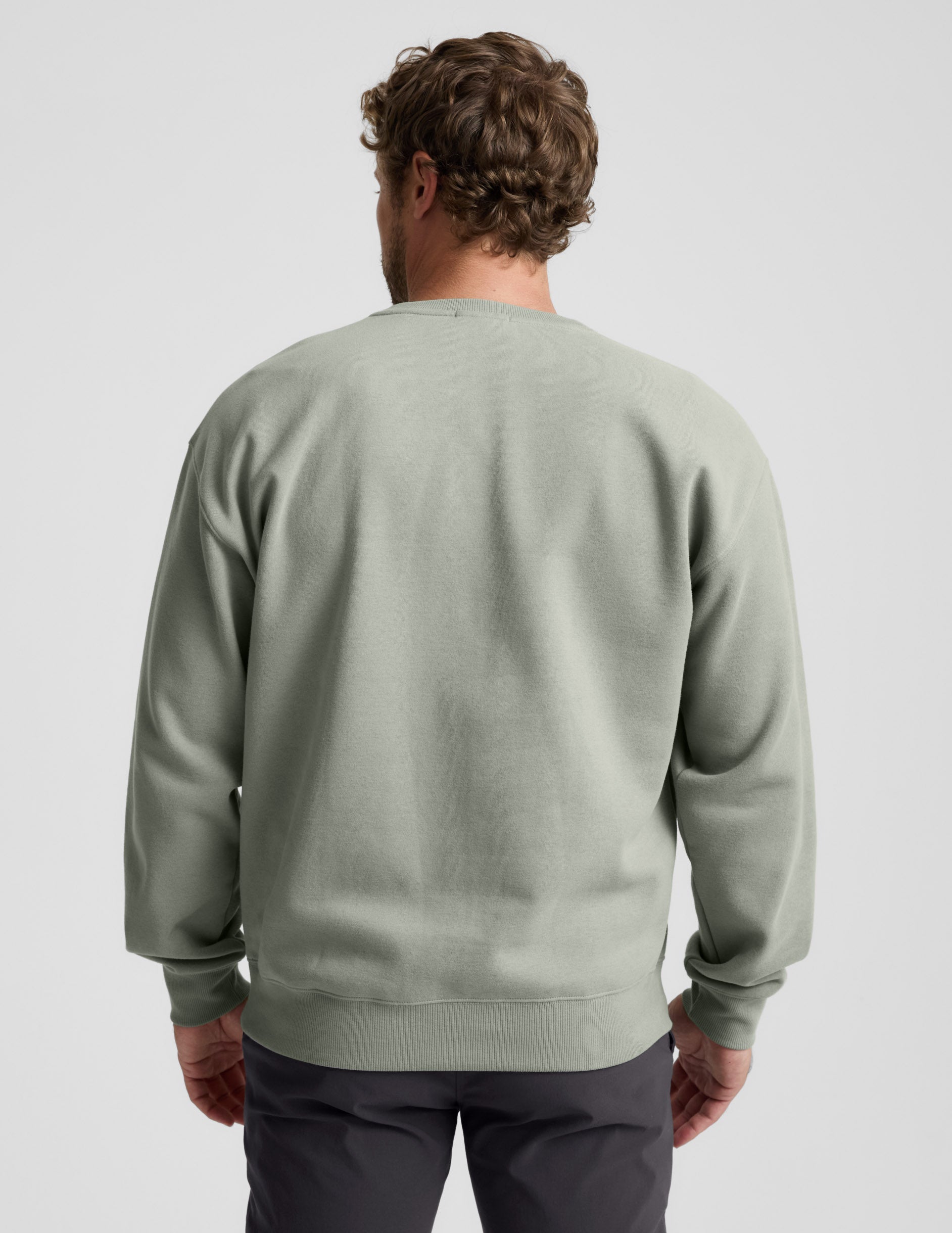 Man wearing a green crewneck sweatshirt and dark pants on a light gray background.