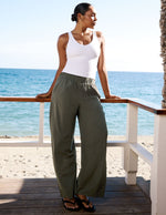 Woman standing on a beach deck wearing a white tank top and green pants.