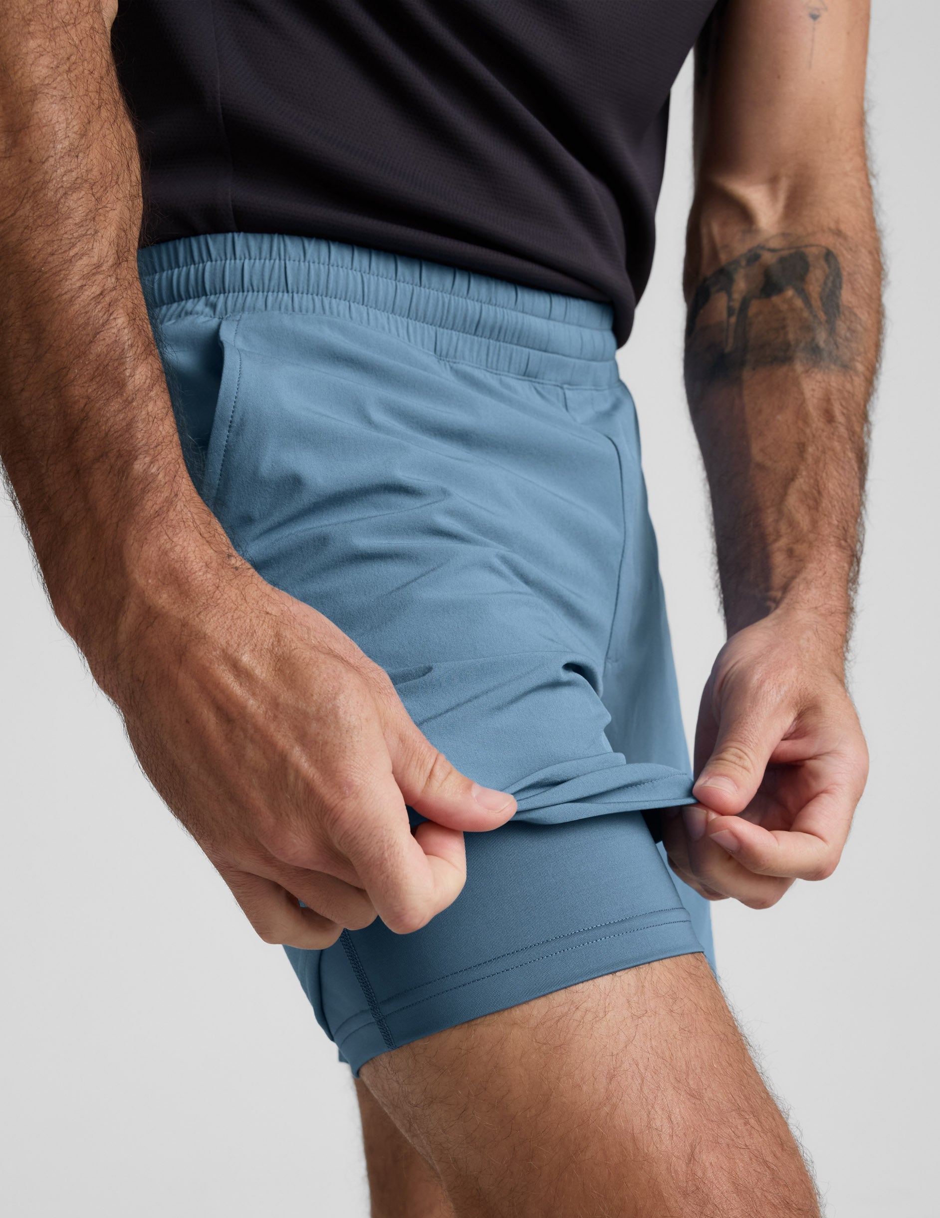 Close-up of blue Pivotal 2.0 Men's Lined Short 7" with a hand lifting the hem to reveal the built-in matching liner.