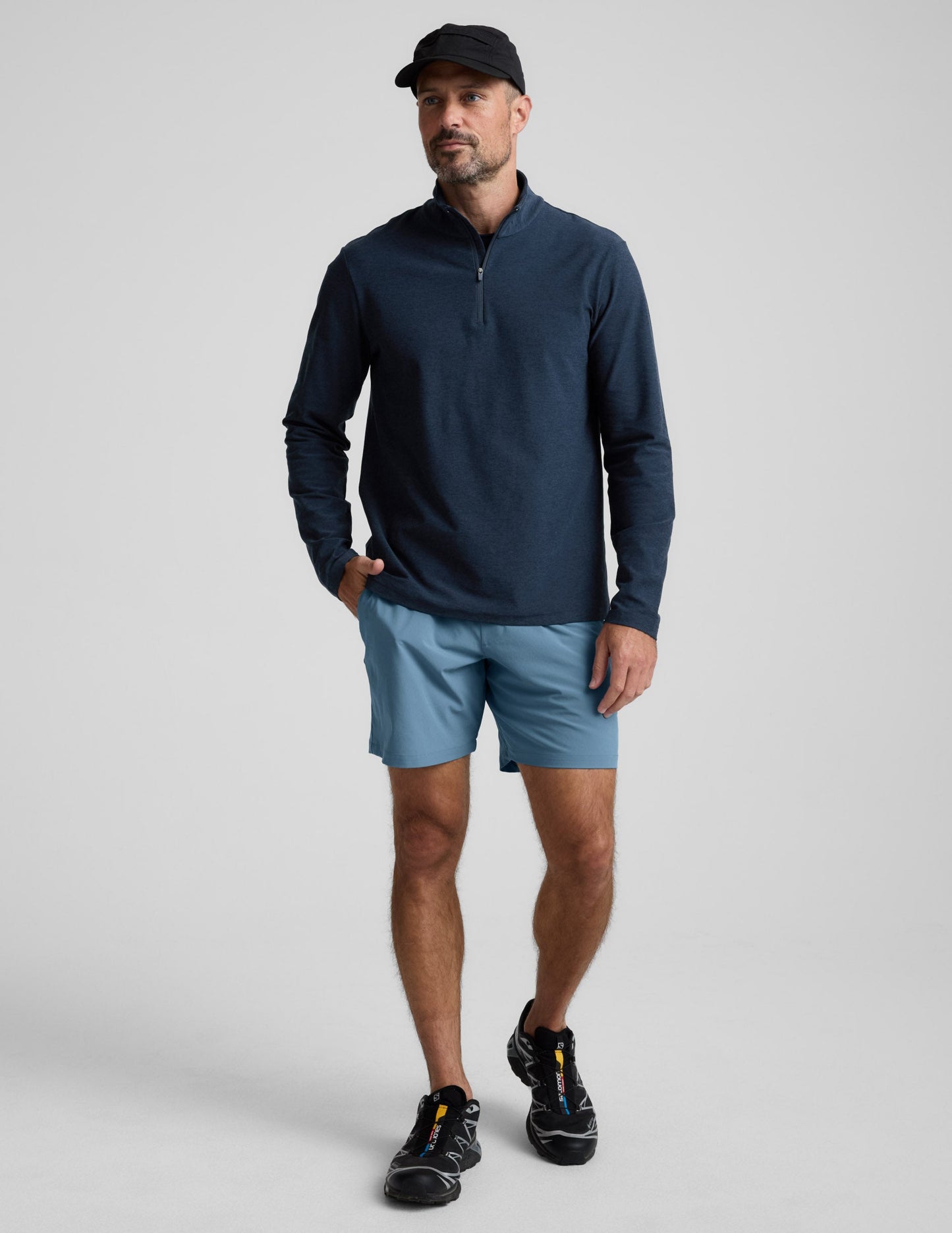 Man wearing Navy Pivotal 2.0 Men's Short 7"™ paired with a navy half-zip pullover, black baseball cap, and black sneakers.