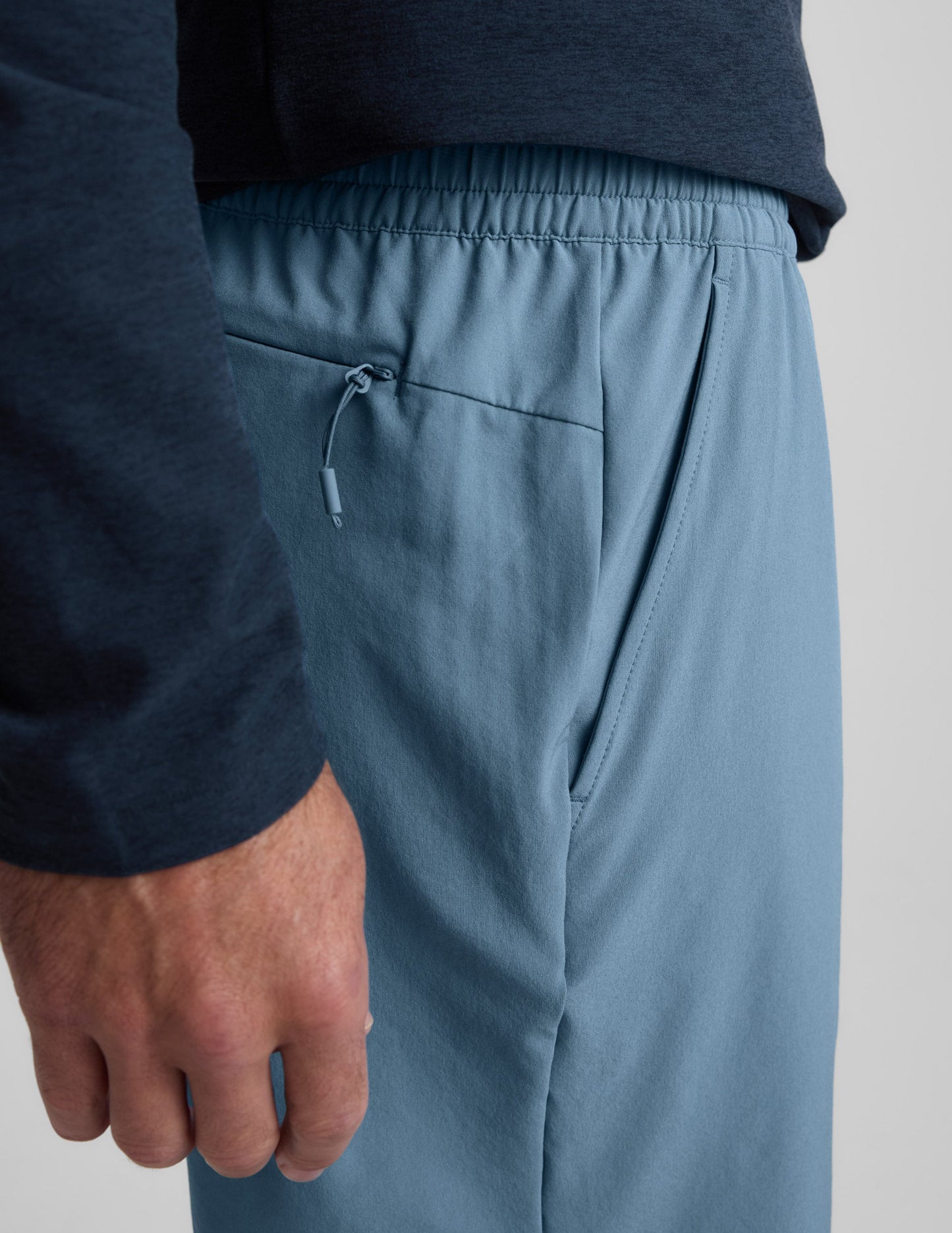 Man wearing Navy Pivotal 2.0 Men's Short 7"™ with a dark long-sleeve shirt, detailing the elastic waist and zipper pocket.