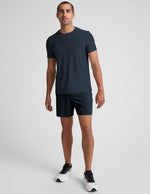 Man stands wearing navy Pivotal 2.0 Men's Short 7", a matching t-shirt, and white sneakers against a gray background.