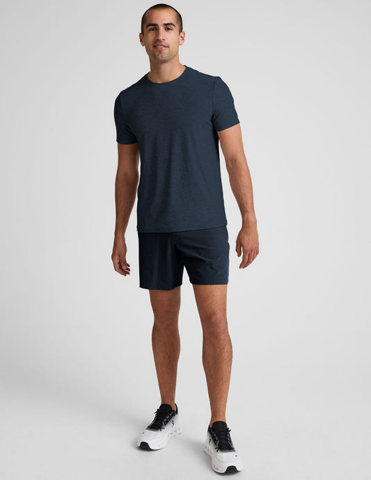 Man stands wearing navy Pivotal 2.0 Men's Short 7", a matching t-shirt, and white sneakers against a gray background.