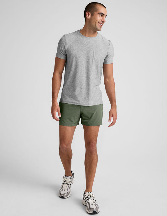 A smiling man walks in olive Pivotal 2.0 Men's Lined Short 5" with a 5-inch inseam, grey tee, and white sneakers.