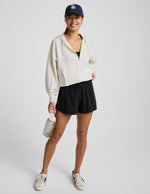 Woman wearing a white zip-up hoodie and black shorts against a plain background.
