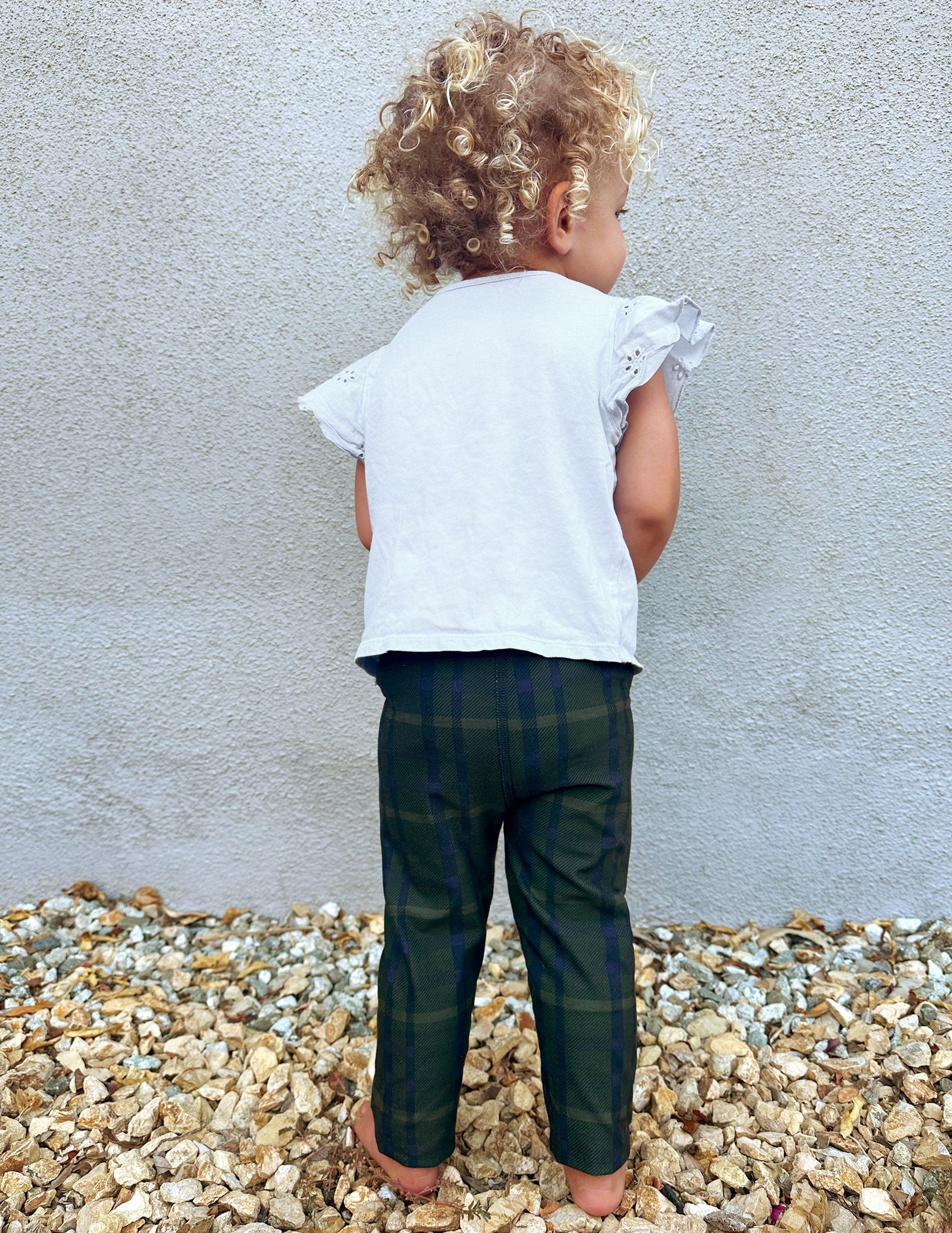 Back view of a toddler wearing Dark Olive Preppy Plaid SoftMark Toddler Leggings, standing on gravel against a gray wall.