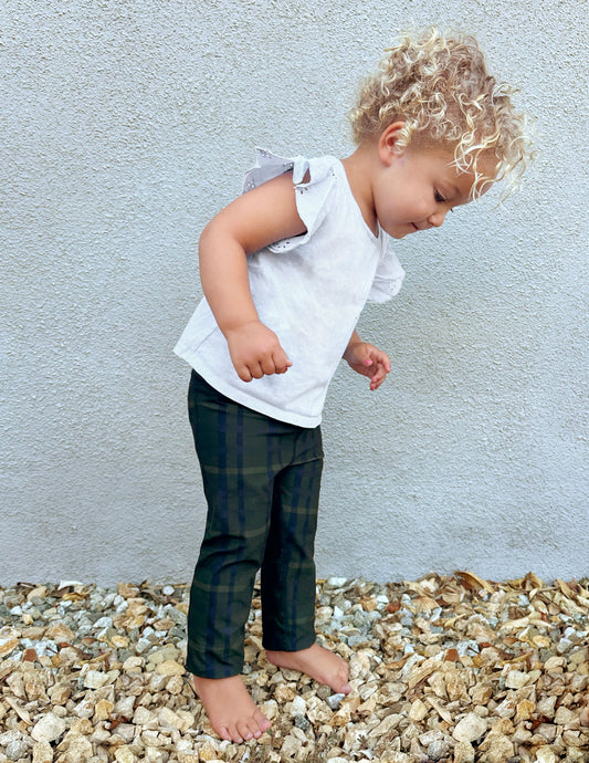 Toddler in Dark Olive Preppy Plaid SoftMark Leggings and white ruffle top standing outdoors on small pebbles.