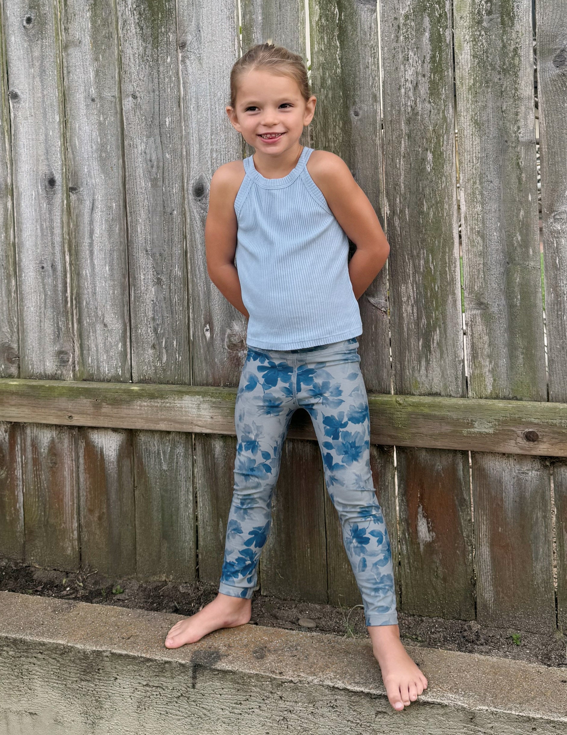 Daydream Meadow SoftMark Toddler Legging with blue floral print, worn by a smiling girl against a wooden fence.
