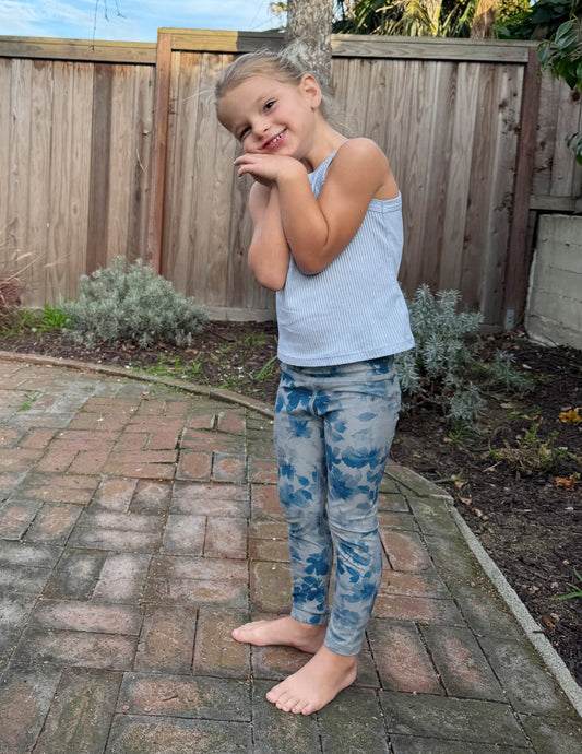 A smiling toddler wears blue floral Daydream Meadow SoftMark Leggings and a blue tank top while standing on a brick patio.