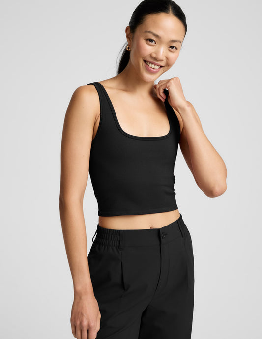 Woman is wearing a black ribbed tank and pants against a light gray background.