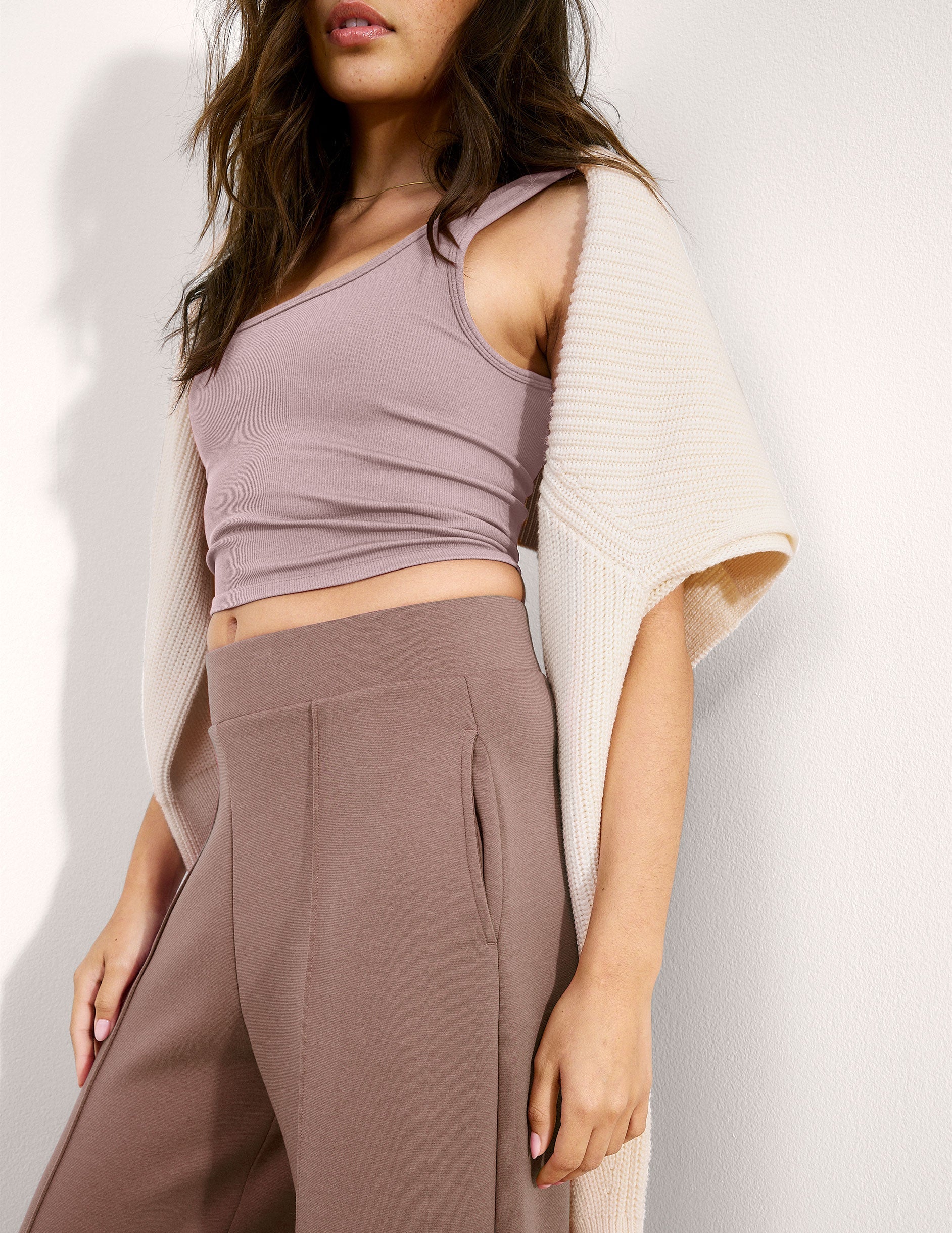 Woman wearing a pink ribbed cropped tank and brown pants against a plain background.