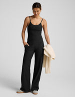 Woman wearing a black sleeveless top and black pants holding a beige cardigan on a light gray background.