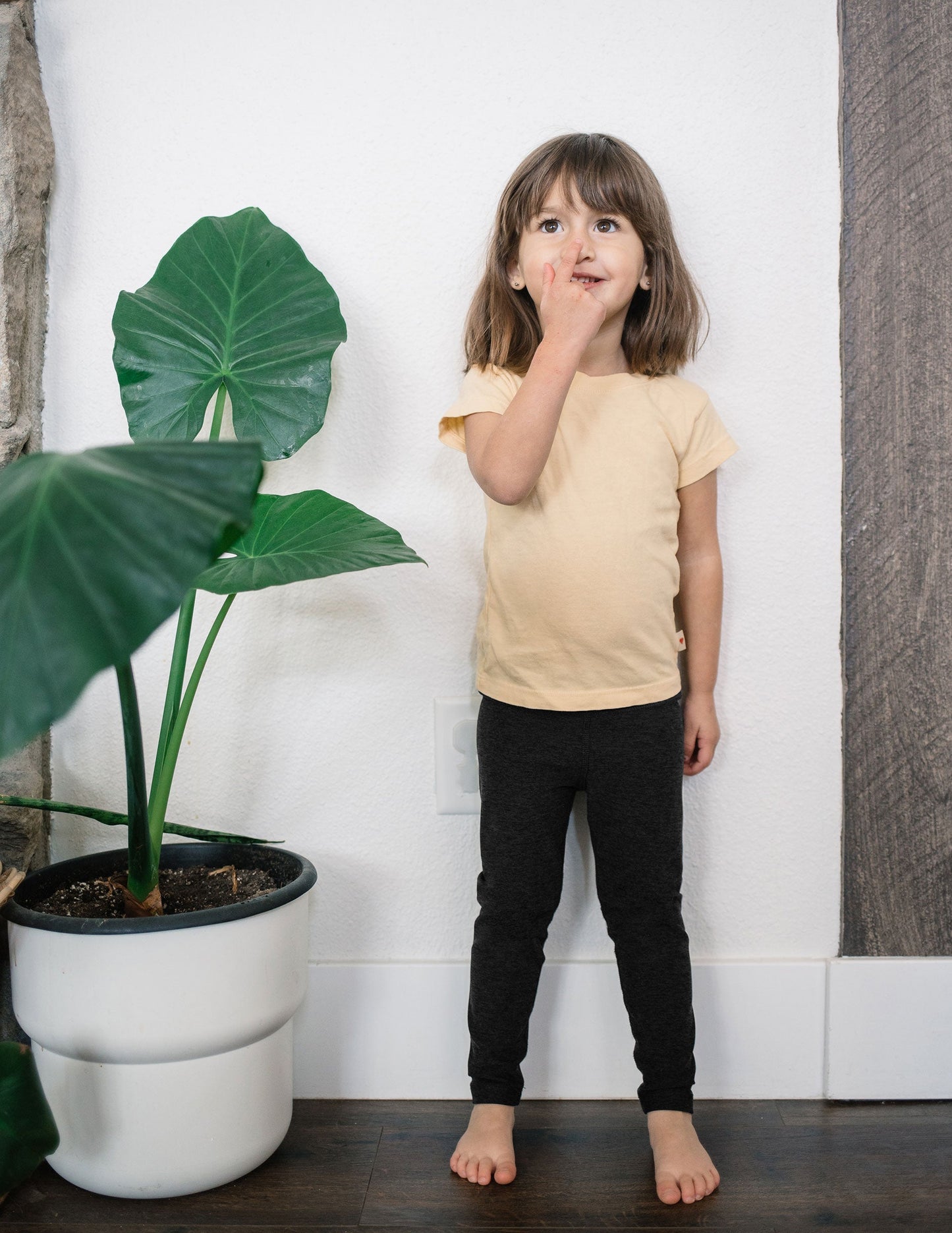 A toddler girl wearing black marled Spacedye Toddler Legging and a yellow t-shirt, standing beside a large potted plant.