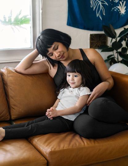 A toddler in black marled Spacedye Toddler Leggings sits on a leather couch with a woman in matching dark activewear.