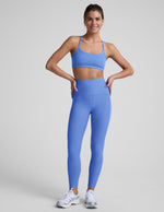 Woman wearing a blue sports bra and blue midi leggings with pockets against a light gray background.