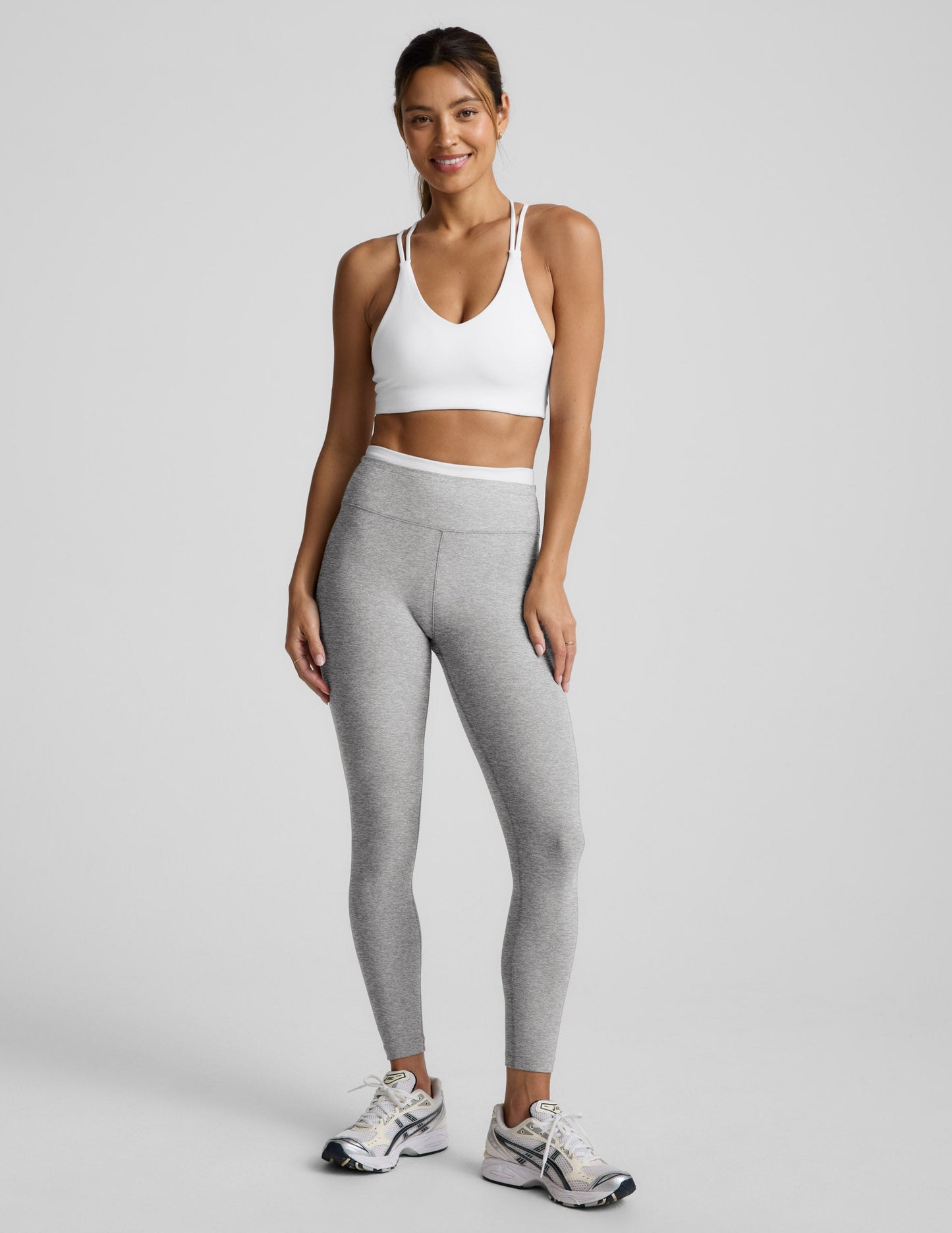 Woman wearing a white sports bra and gray leggings on a light gray background.