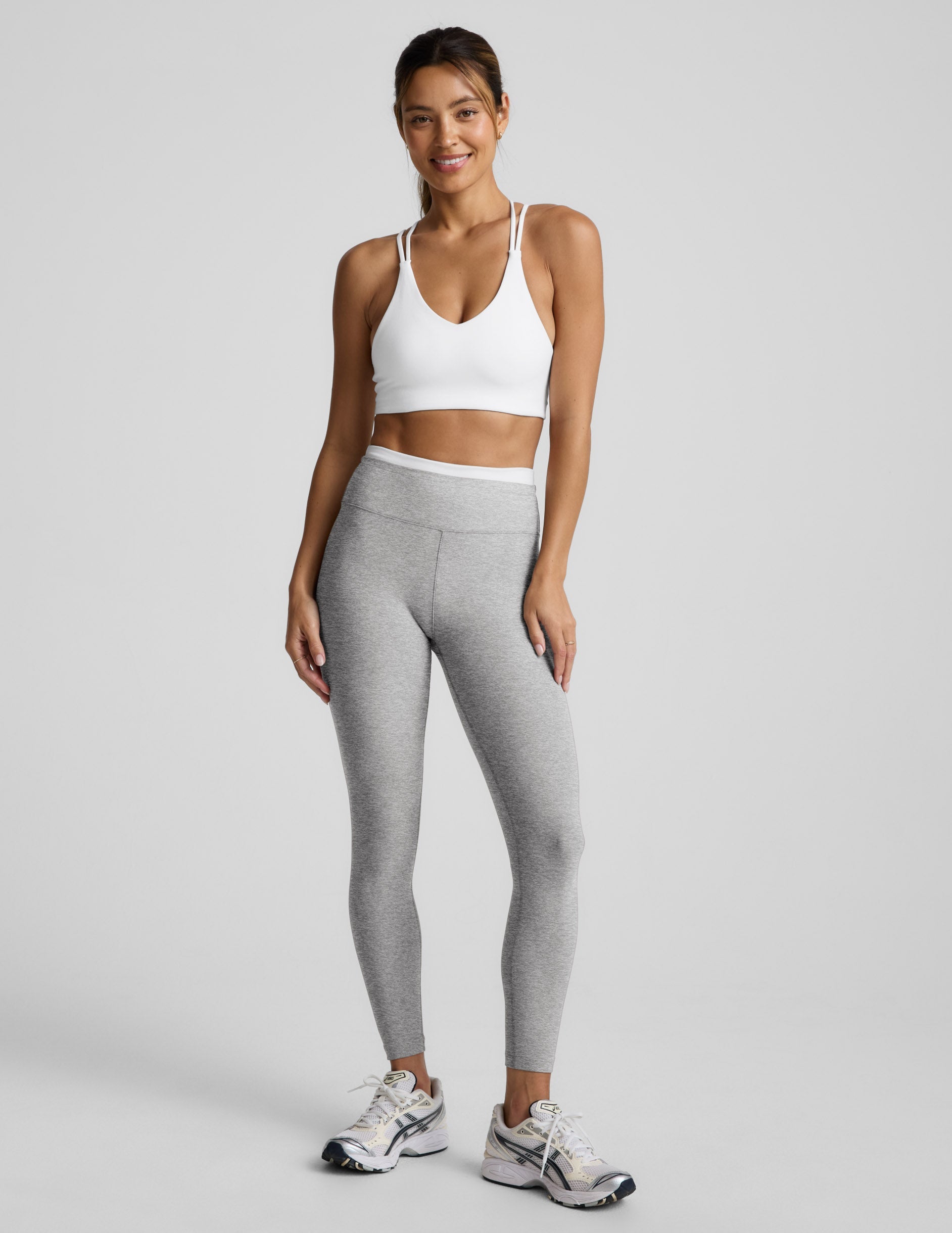 Woman wearing a white sports bra and gray leggings on a light gray background.