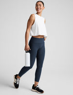 Woman wearing a white sleeveless top and dark blue leggings, holding a white water bottle on a light gray background.