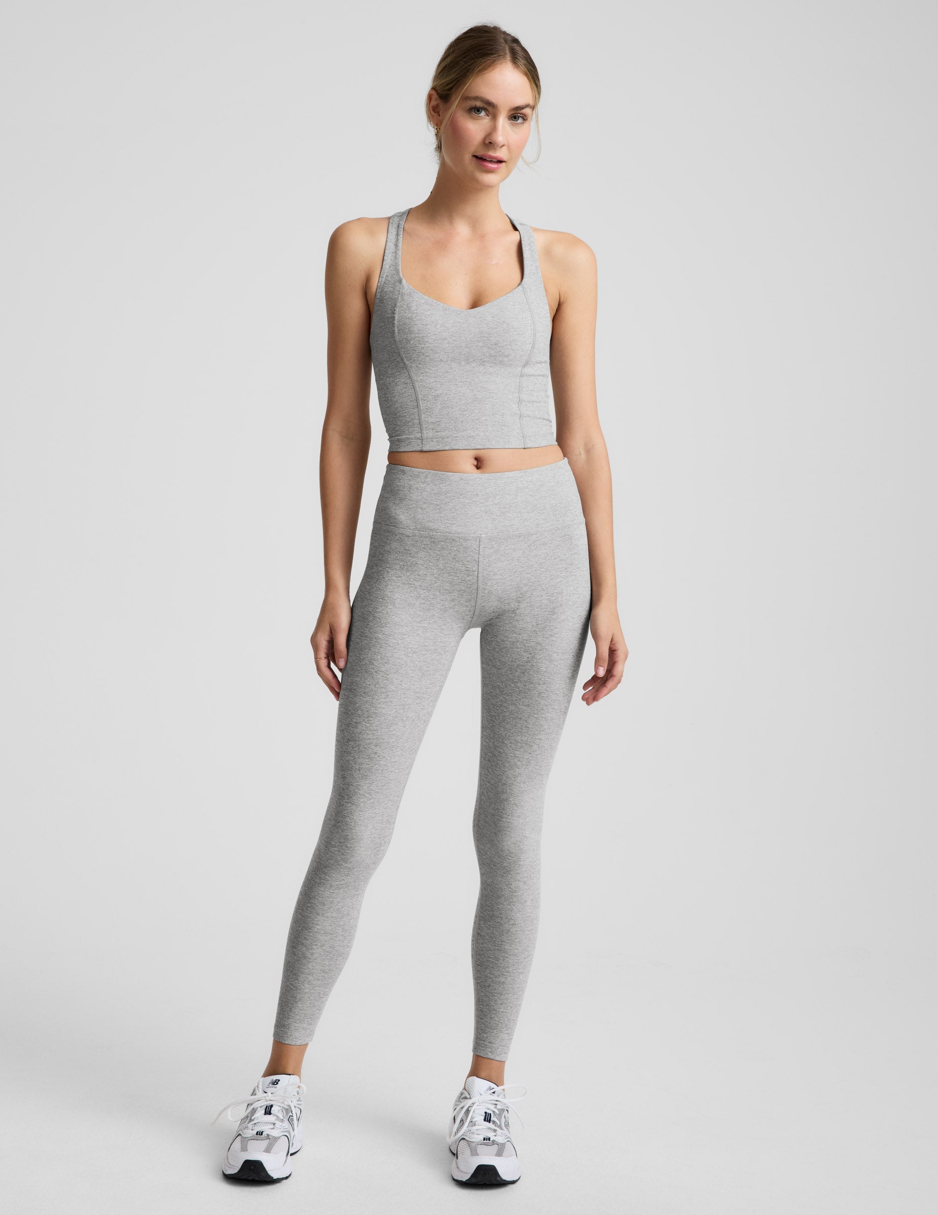 Woman wearing a silver mist cropped racerback tank and mid rise midi length leggings against a plain background.
