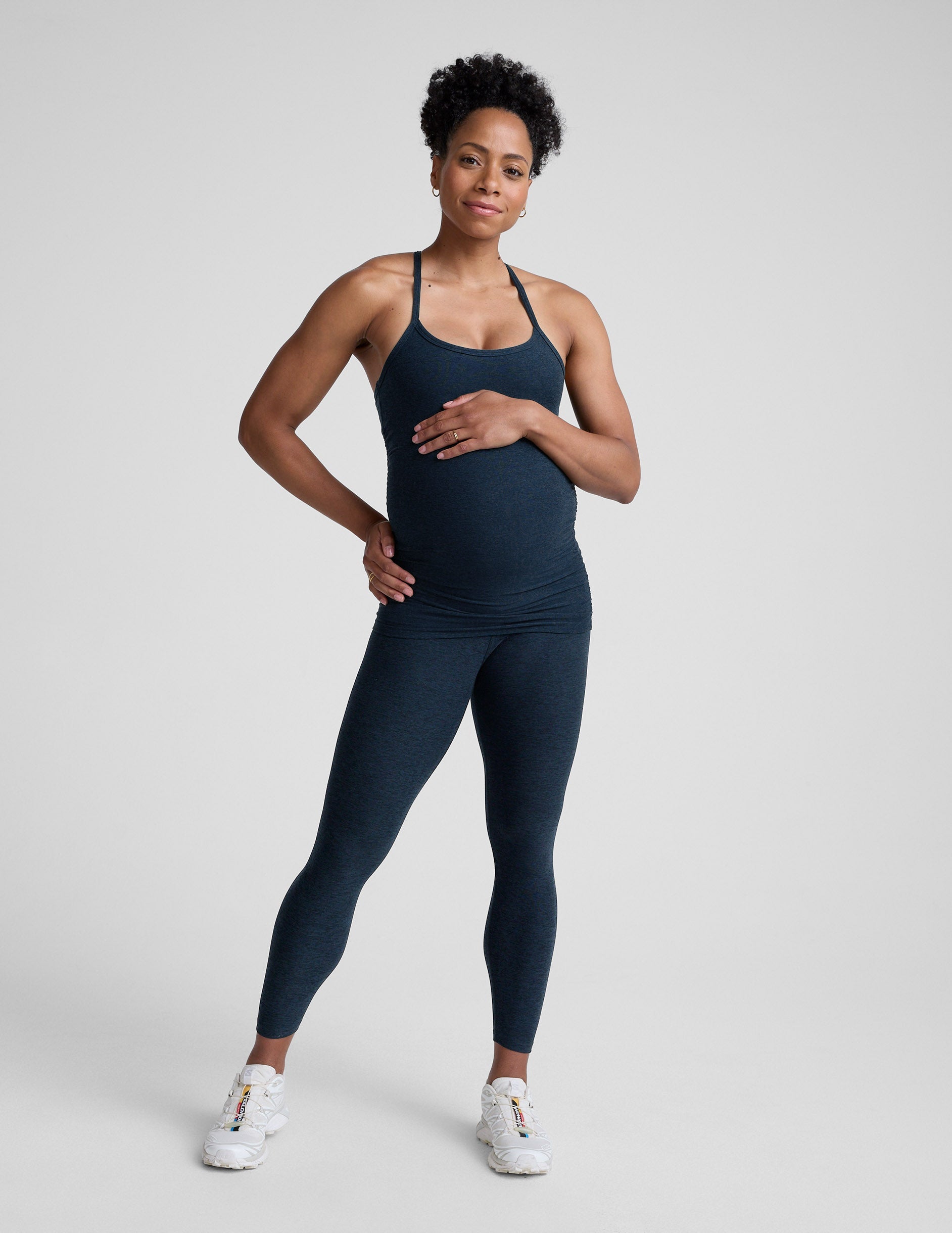 Pregnant woman wearing a dark blue tank top and leggings on a light gray background.