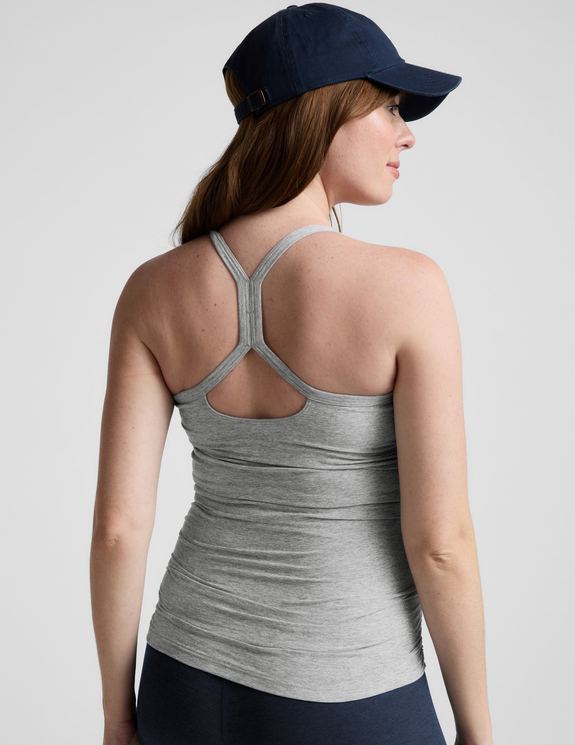 Woman wearing a gray maternity tank top and navy cap on a light gray background.