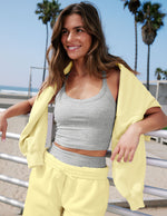 Woman in gray tank top and yellow fleece standing on a beach with palm trees in the background.