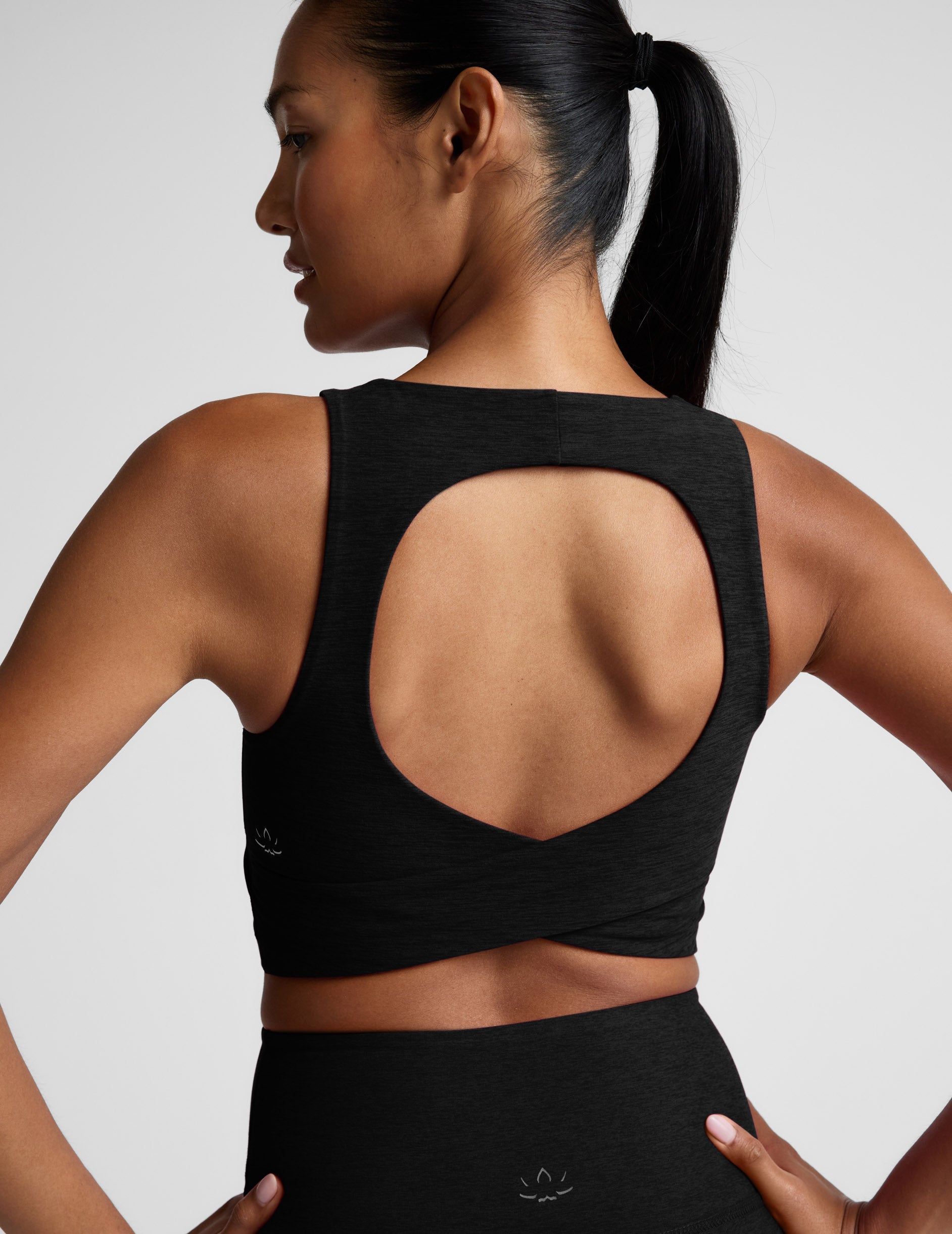 Woman in a black cropped tank with a crossover strap in the back and black leggings against a light gray background.