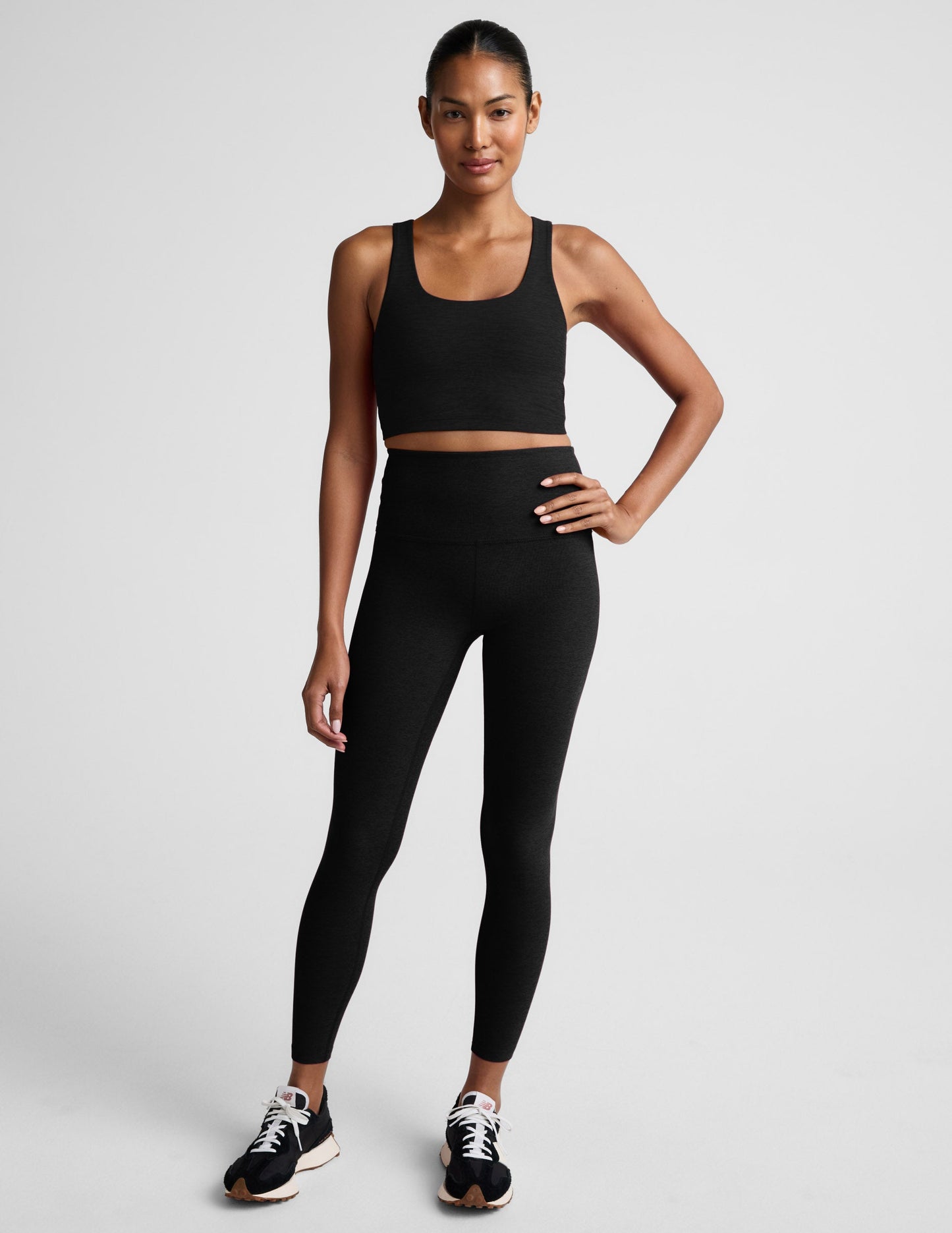 Woman in a black cropped tank with a crossover strap in the back and black leggings against a light gray background.
