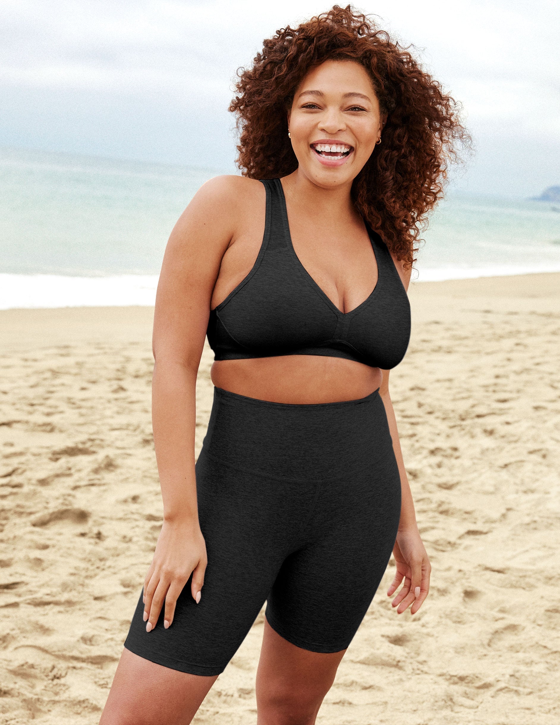 Smiling woman on a beach in black marled Spacedye High Waisted 7" Biker Shorts and a matching sports bra.