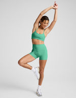 Woman in yoga pose wearing green Spacedye Keep Pace 3" Short with high waistband and matching top, arms raised, smiling.
