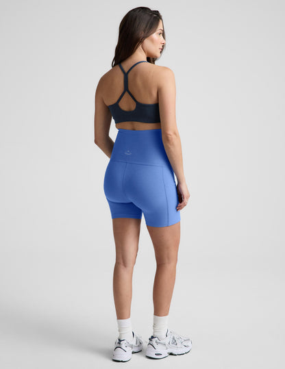 Rear view of blue Spacedye Keep Pace 5" Maternity Pocket Biker Shorts on a model, showing high waist and side pocket.