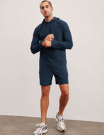 A man stands wearing navy Spacedye Unlimited Men's Shorts with a matching hoodie and white athletic sneakers.