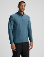 Man wearing a blue long-sleeve with a quarter zip and black pants on a light gray background.
