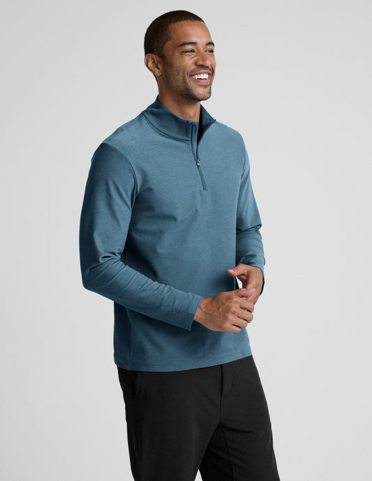 Man wearing a blue long-sleeve with a quarter zip and black pants on a light gray background.