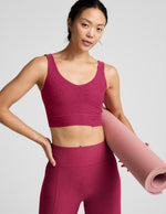 Woman wearing a red long line bra and leggings holding a pink yoga mat on a light gray background.