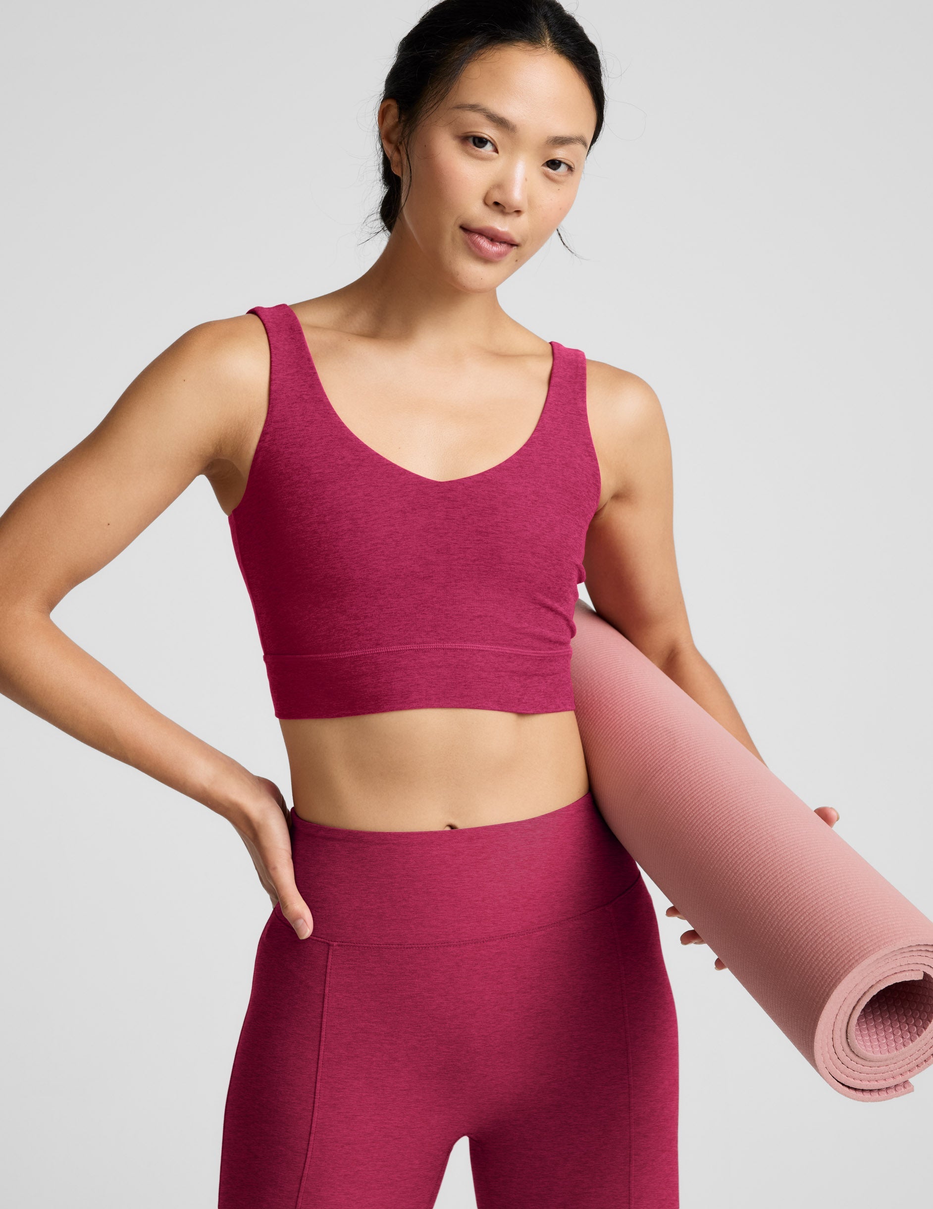 Woman wearing a red long line bra and leggings holding a pink yoga mat on a light gray background.