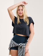 Woman wearing a blue crop top and dark shorts with a striped sweater around her waist on a plain background.