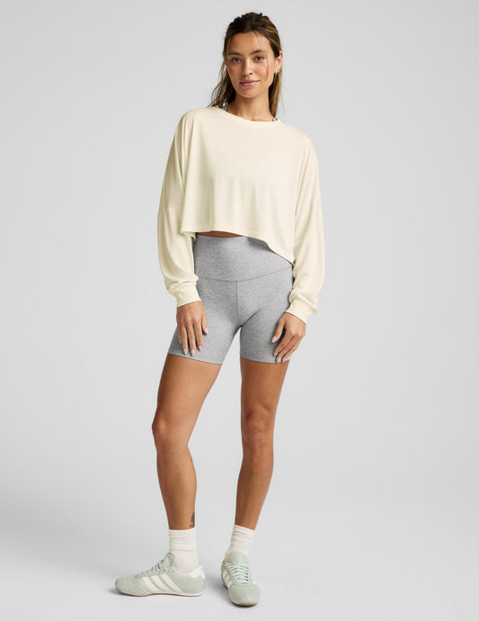 Woman wearing a beige long-sleeve top and gray shorts on a light gray background.