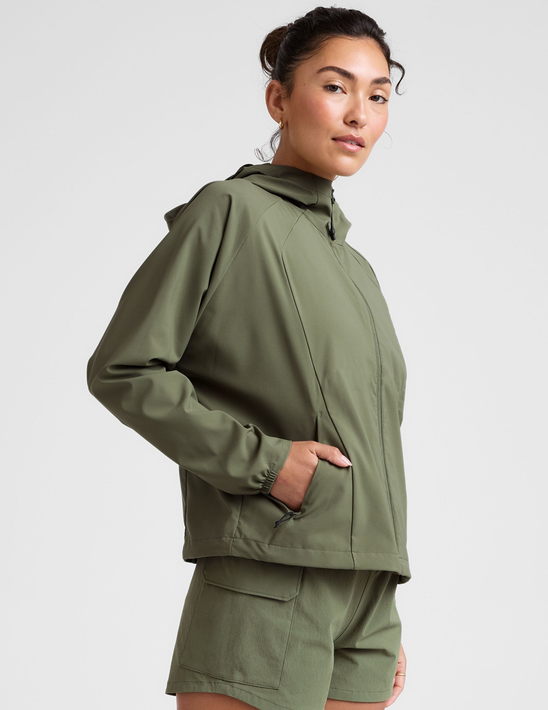 model is wearing an olive green zip-up jacket with side pockets and a hood on a white background.